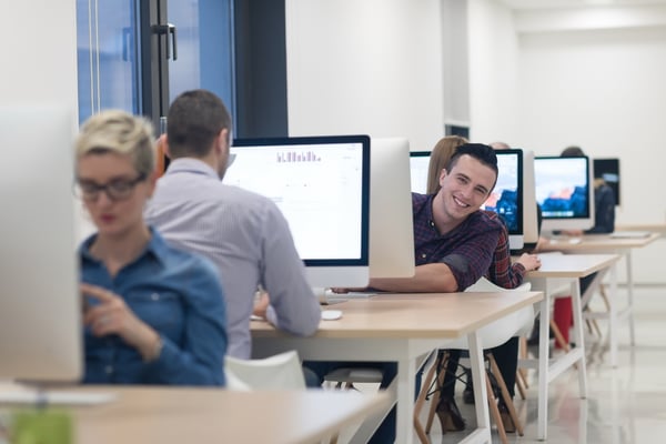 man leaning round his monitor to smile at his colleague